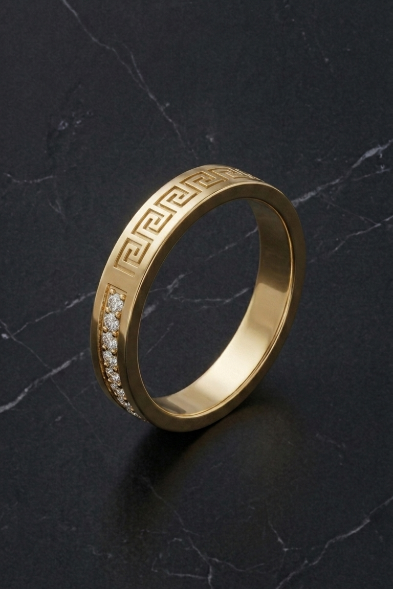 A gold band ring featuring a Greek key pattern and a row of small clear stones on one side, displayed on a dark marble surface.