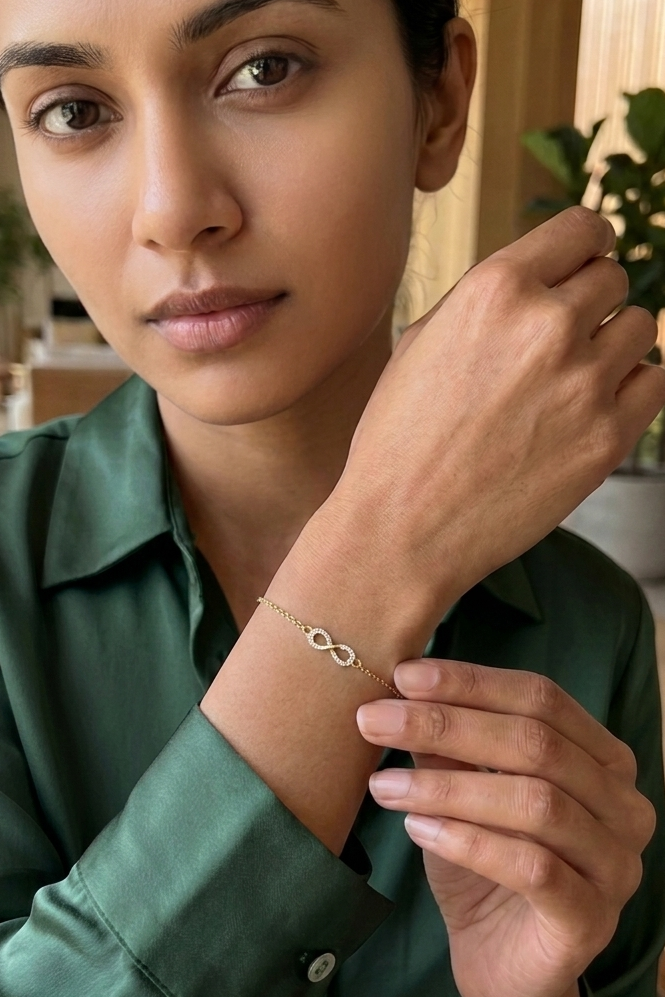 A woman wearing a delicate gold and diamond infinity bracelet, shown in a close-up shot. She is wearing a dark green satin shirt, and the background is a softly blurred indoor setting with warm lighting