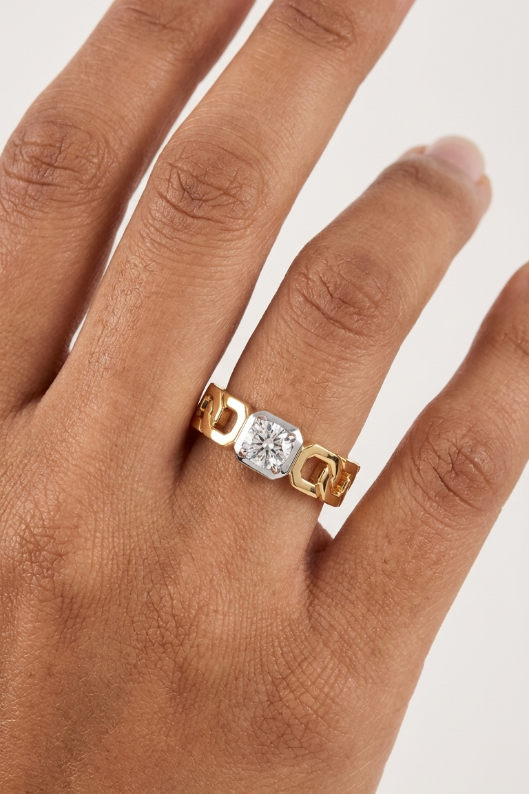 A close-up of a hand wearing a gold ring with a cushion-cut clear gemstone in a bezel setting, photographed against a light neutral background.