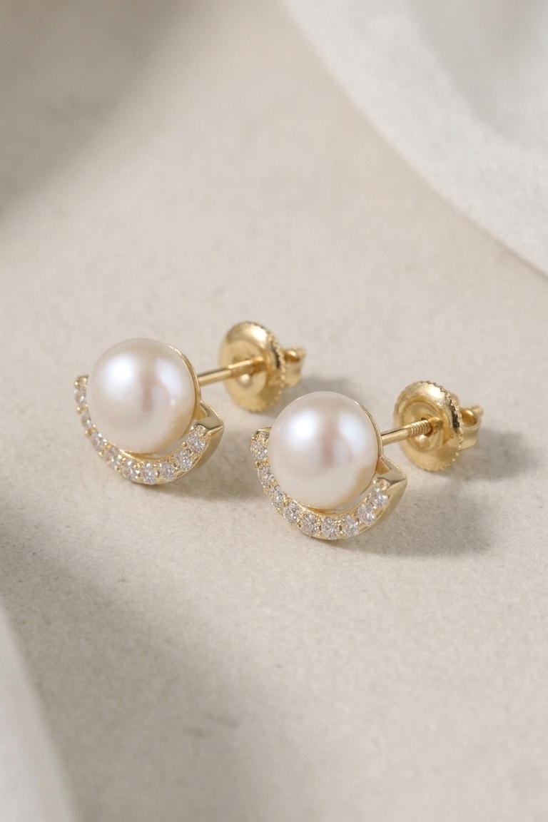 A pair of gold crescent-shaped stud earrings with white freshwater pearls and small clear crystal accents, displayed on a soft beige textured surface.