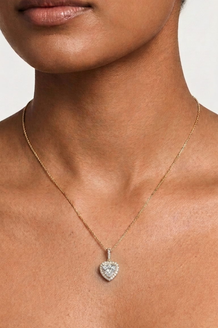 A close-up of a model wearing a delicate gold chain necklace with a heart-shaped pendant paved with small clear stones.