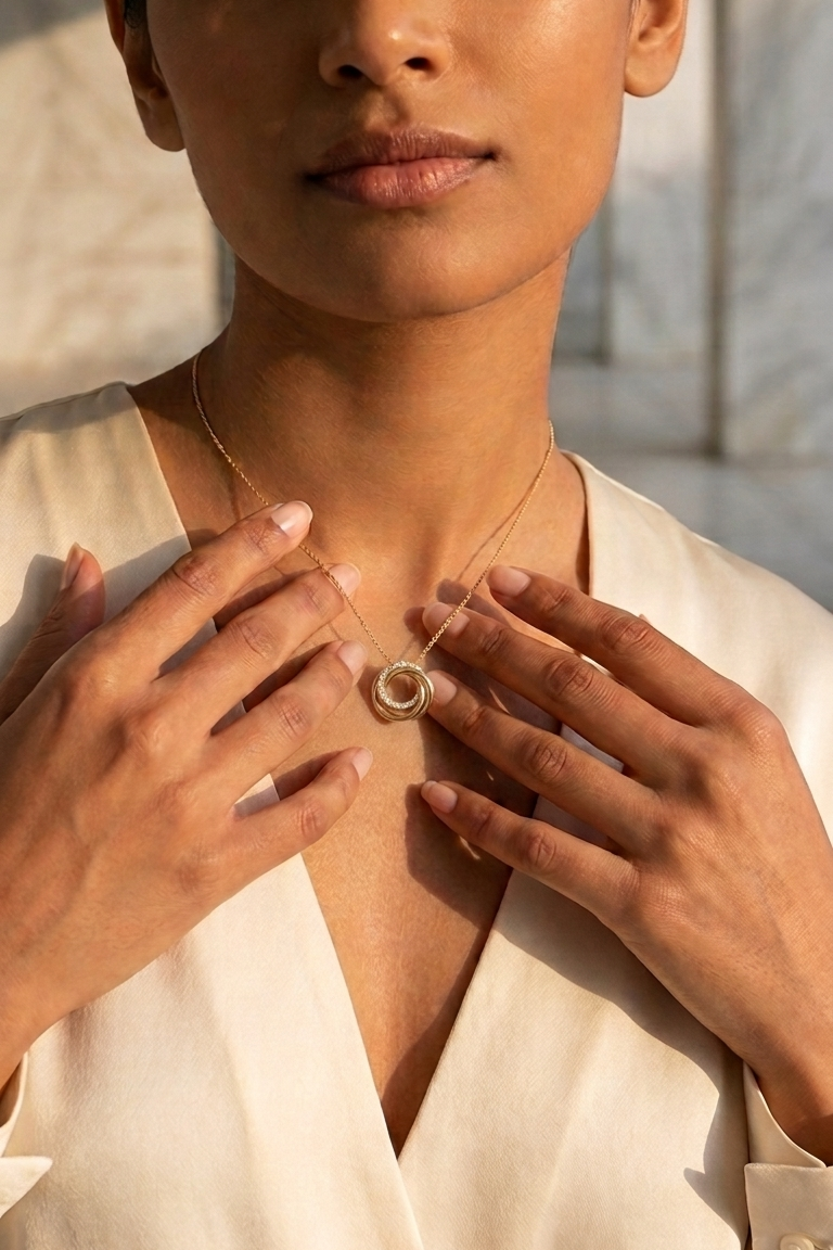 A close-up lifestyle shot of a woman wearing a delicate gold necklace with a circular pendant, touching the necklace with both hands. She is wearing a cream-colored top, and the background is a softly blurred stone wall with warm sunlight.